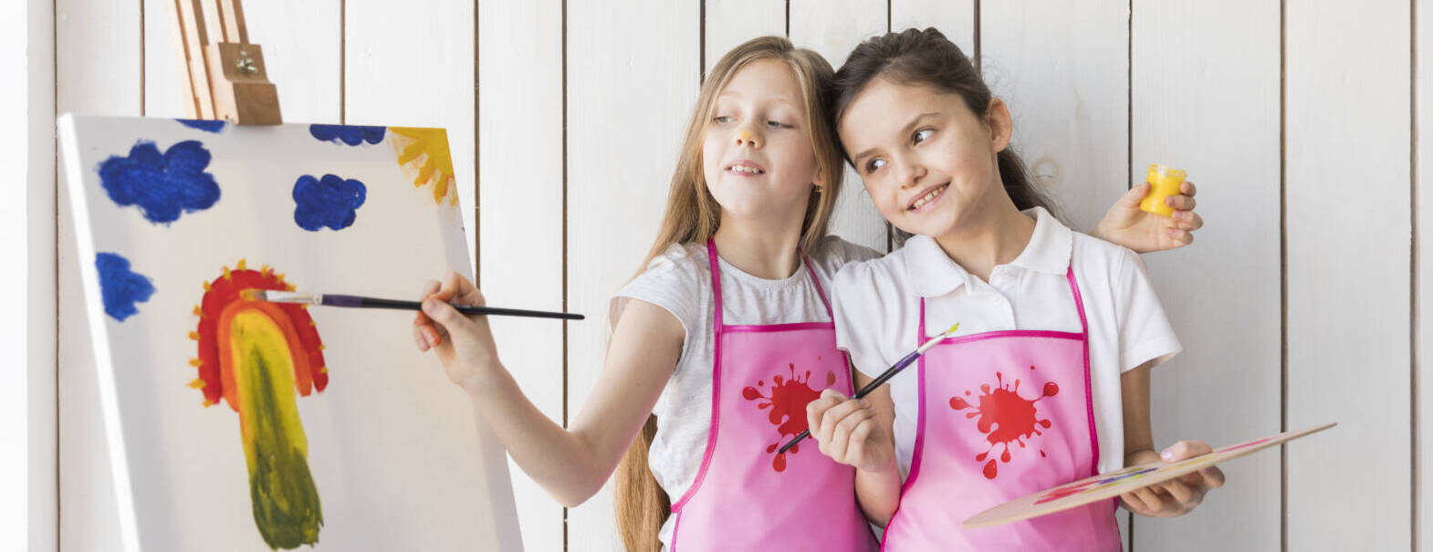 happy-girl-holding-palette-hand-looking-her-friend-painting-canvas-with-brush